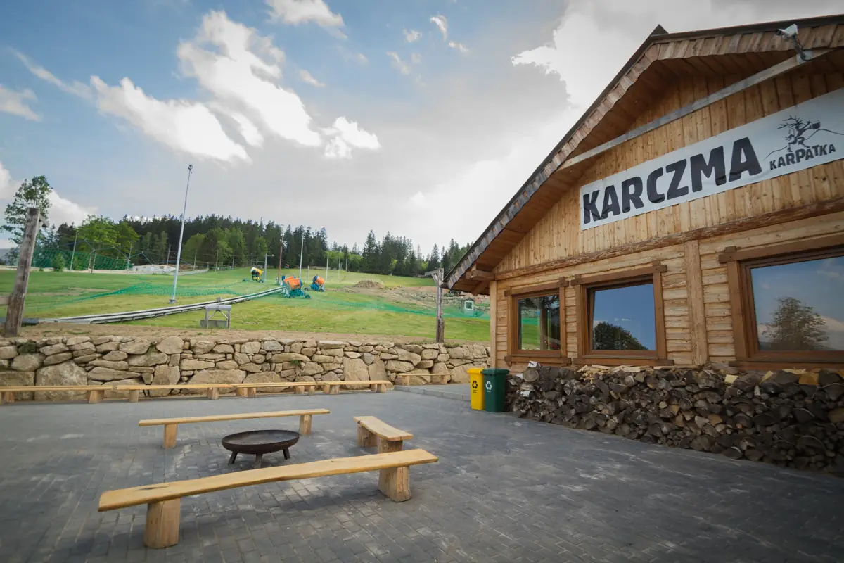 Karczma Karpatka latem — drewniany budynek z paleniskiem i ławkami u podnóża stoku w Karpaczu